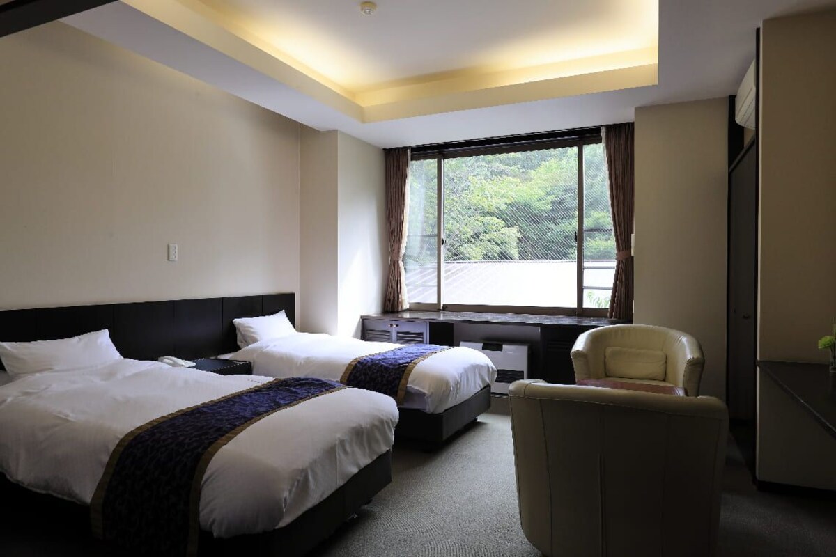 A hotel room with two beds, a desk, a chair, and a window with a view of greenery.