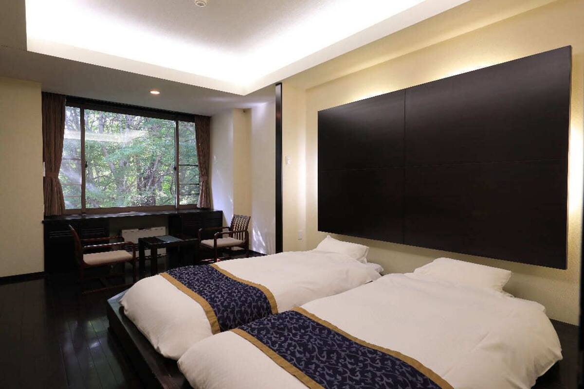 A hotel room with two beds, a desk, and a window with a view of trees.