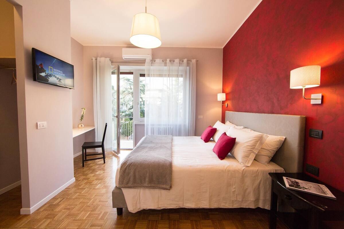 A bedroom with a bed, a desk, a chair, a TV, and a window with curtains.
