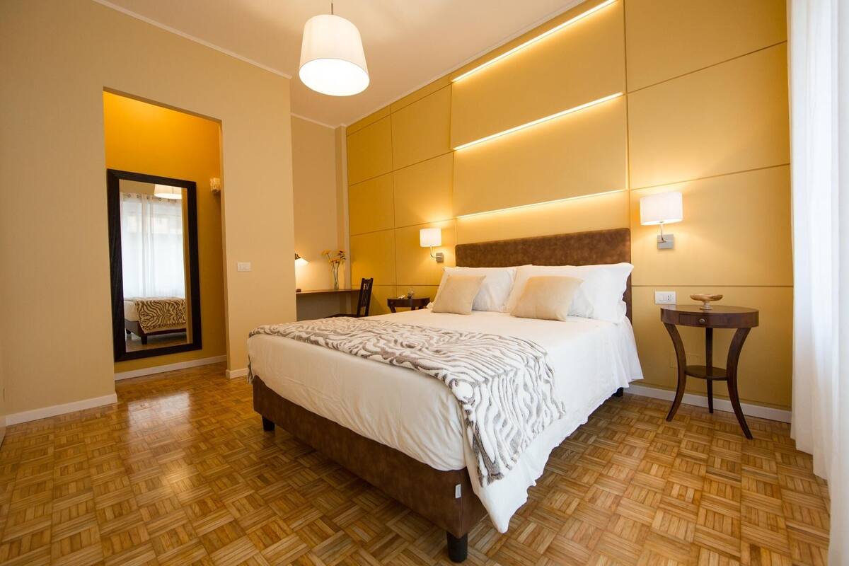 A hotel room with a large bed, wooden flooring, a mirror, a bedside table, and a wall-mounted light fixture.