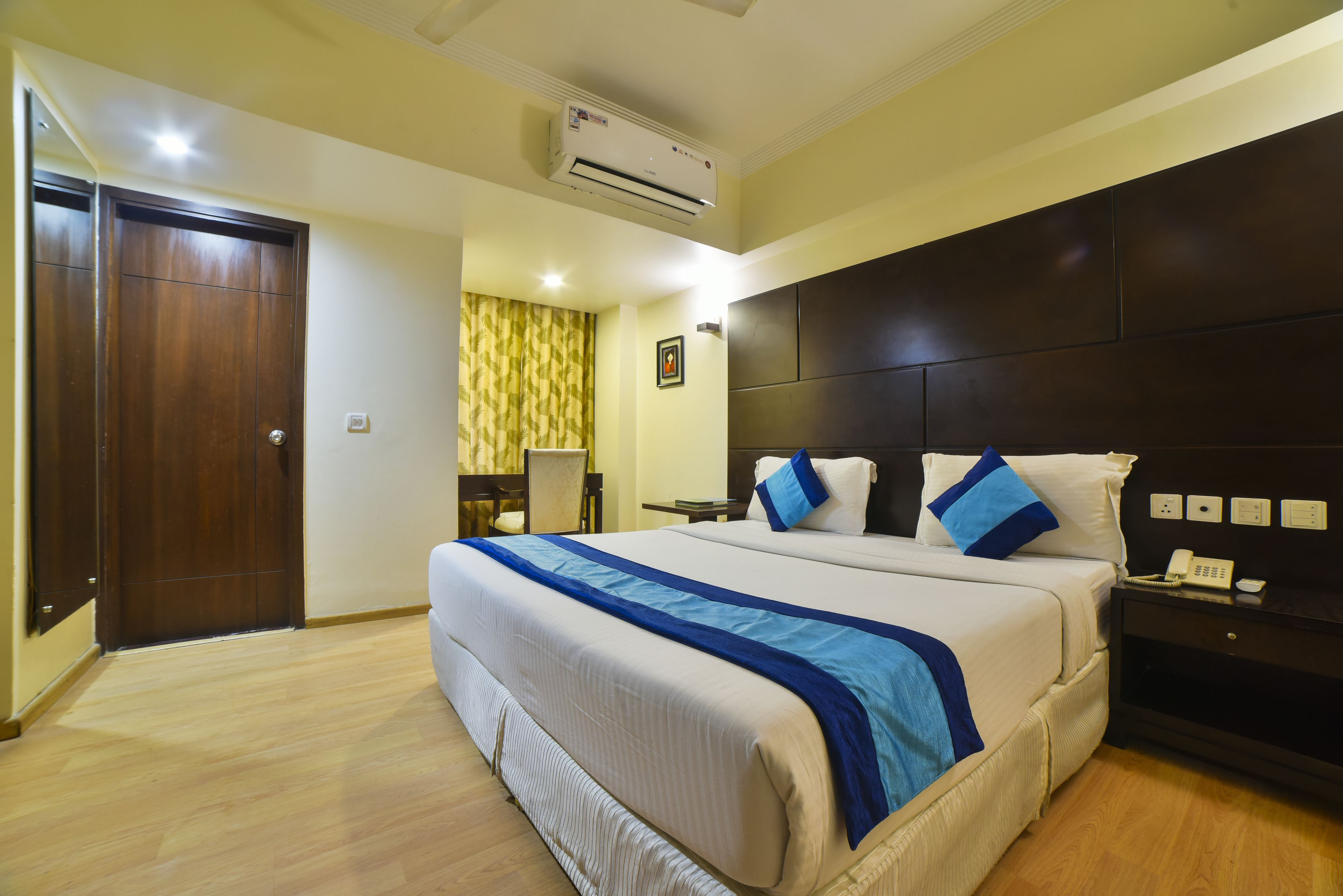 A hotel room with a large bed, a bedside table with a phone, a wall-mounted air conditioner, and a door.