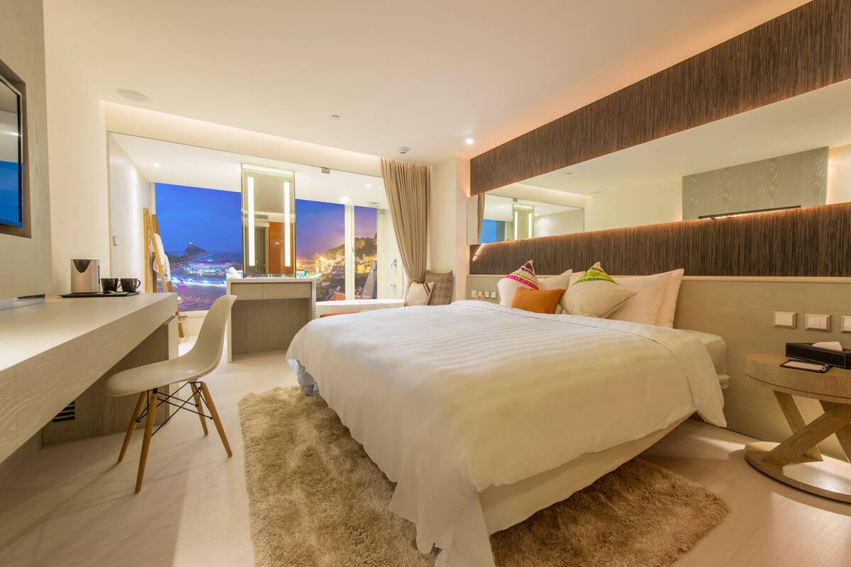 Une chambre d’hôtel moderne, dotée d’un grand lit, d’un bureau et offrant une vue sur le paysage urbain au crépuscule.