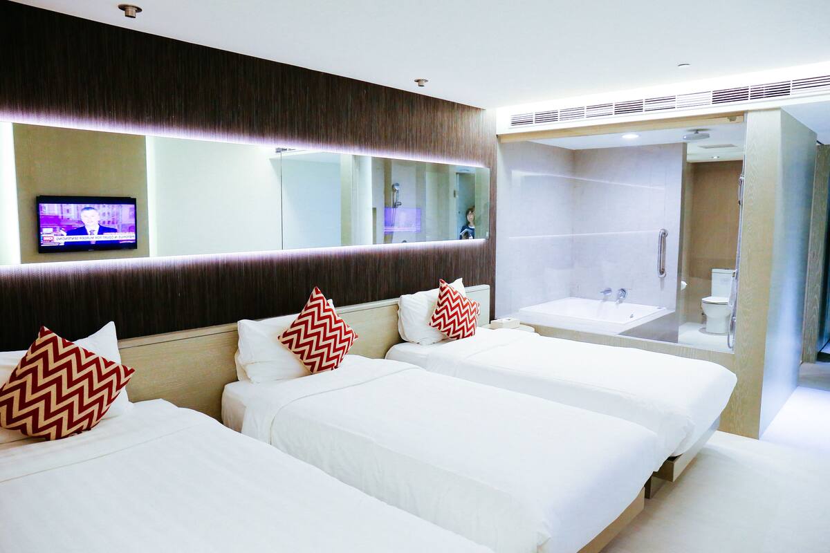 Une chambre d’hôtel avec trois lits, un grand miroir, une télévision et une salle de bain équipée d’un lavabo et d’une douche.