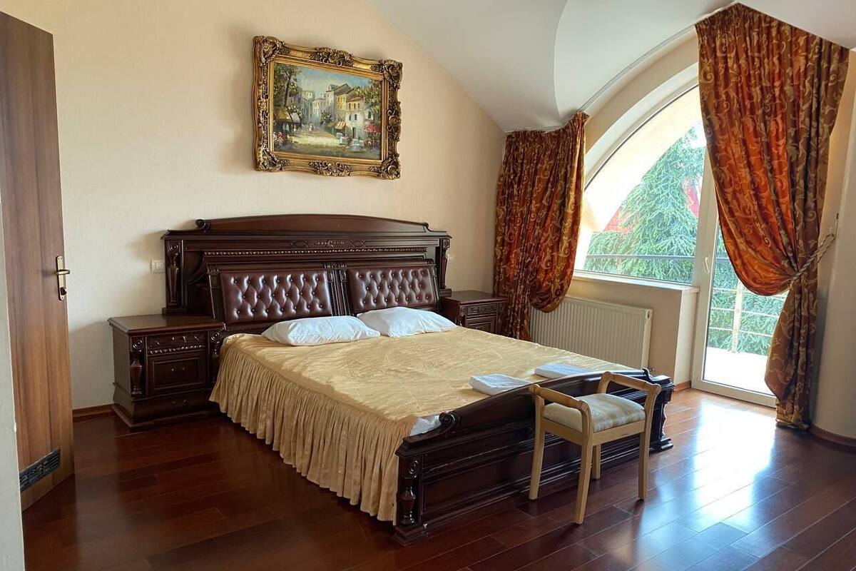 A bedroom with a large bed, a nightstand, a chair, a window with curtains, and a painting on the wall.