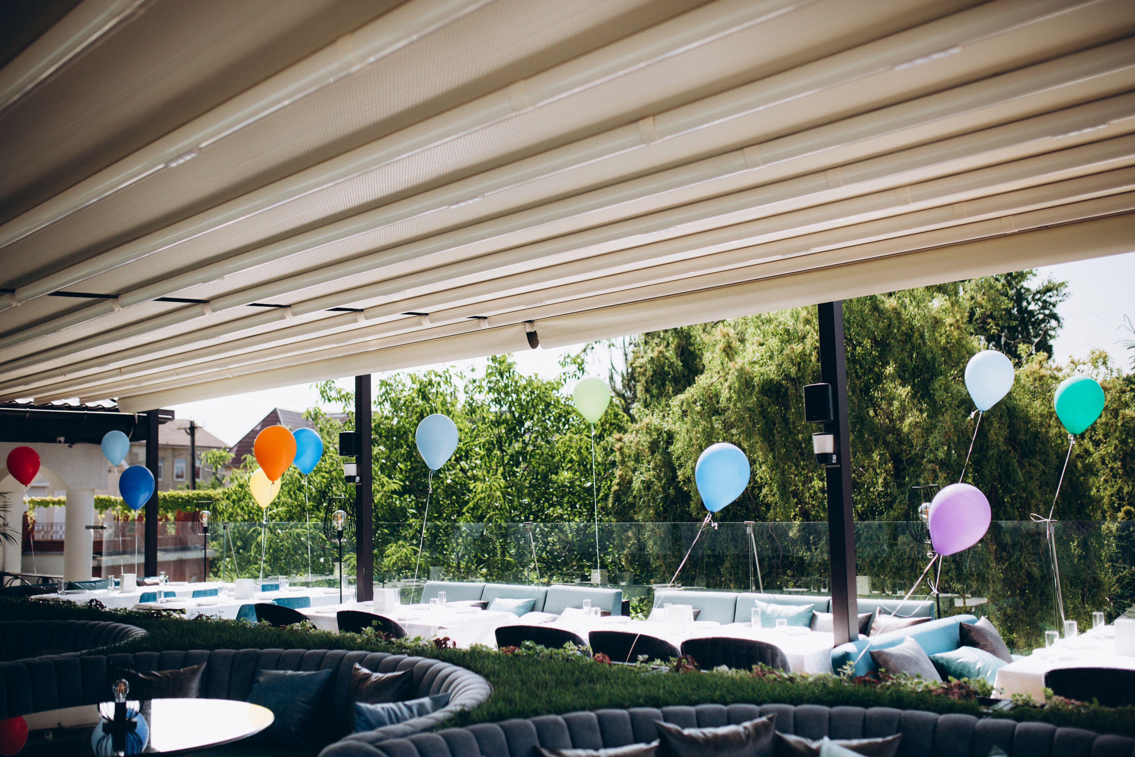 An outdoor seating area with round tables and cushioned benches, surrounded by a covered patio with a view of trees.