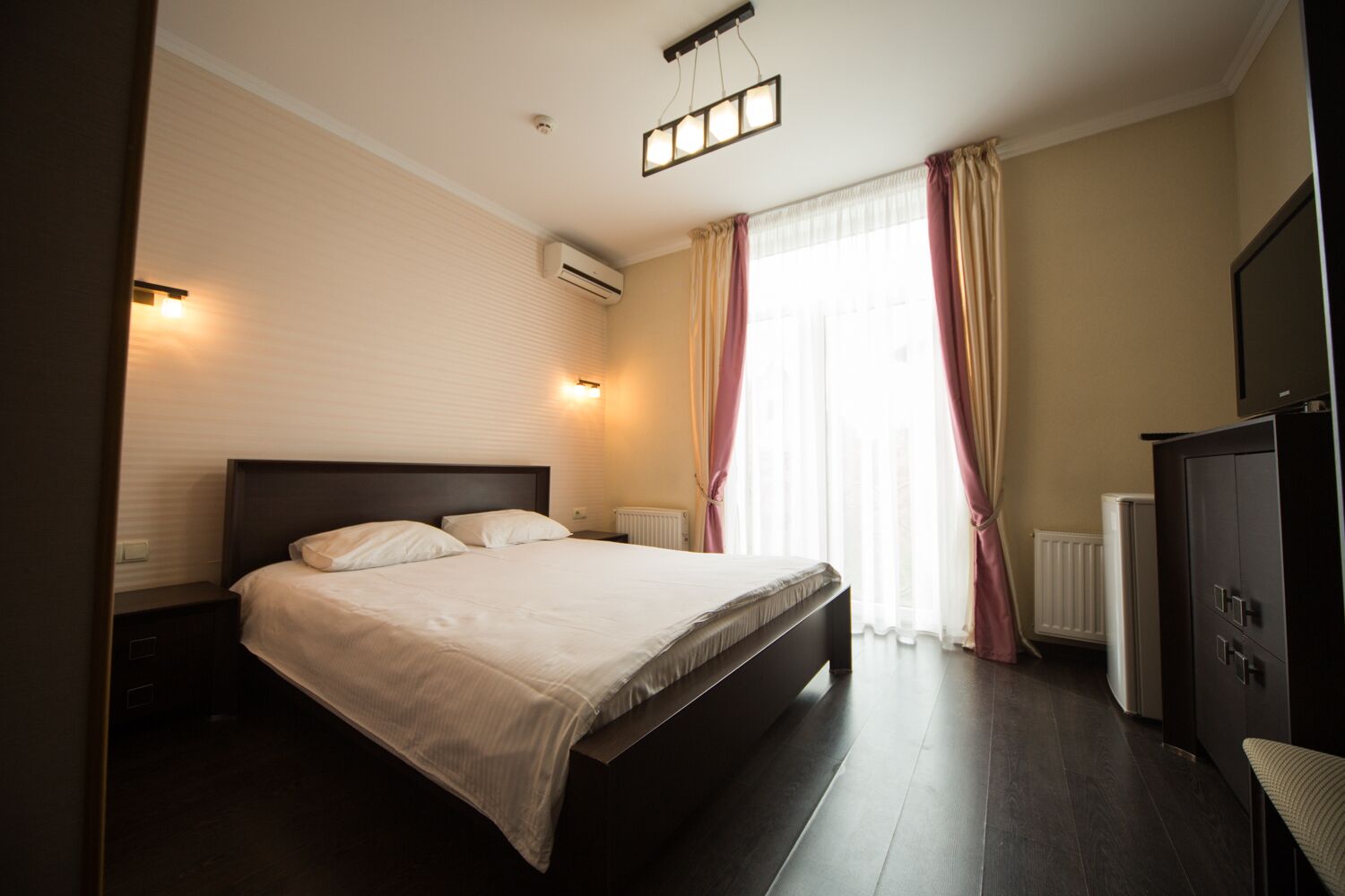 A modern hotel room with a large bed, a radiator, a wardrobe, and a window with curtains.
