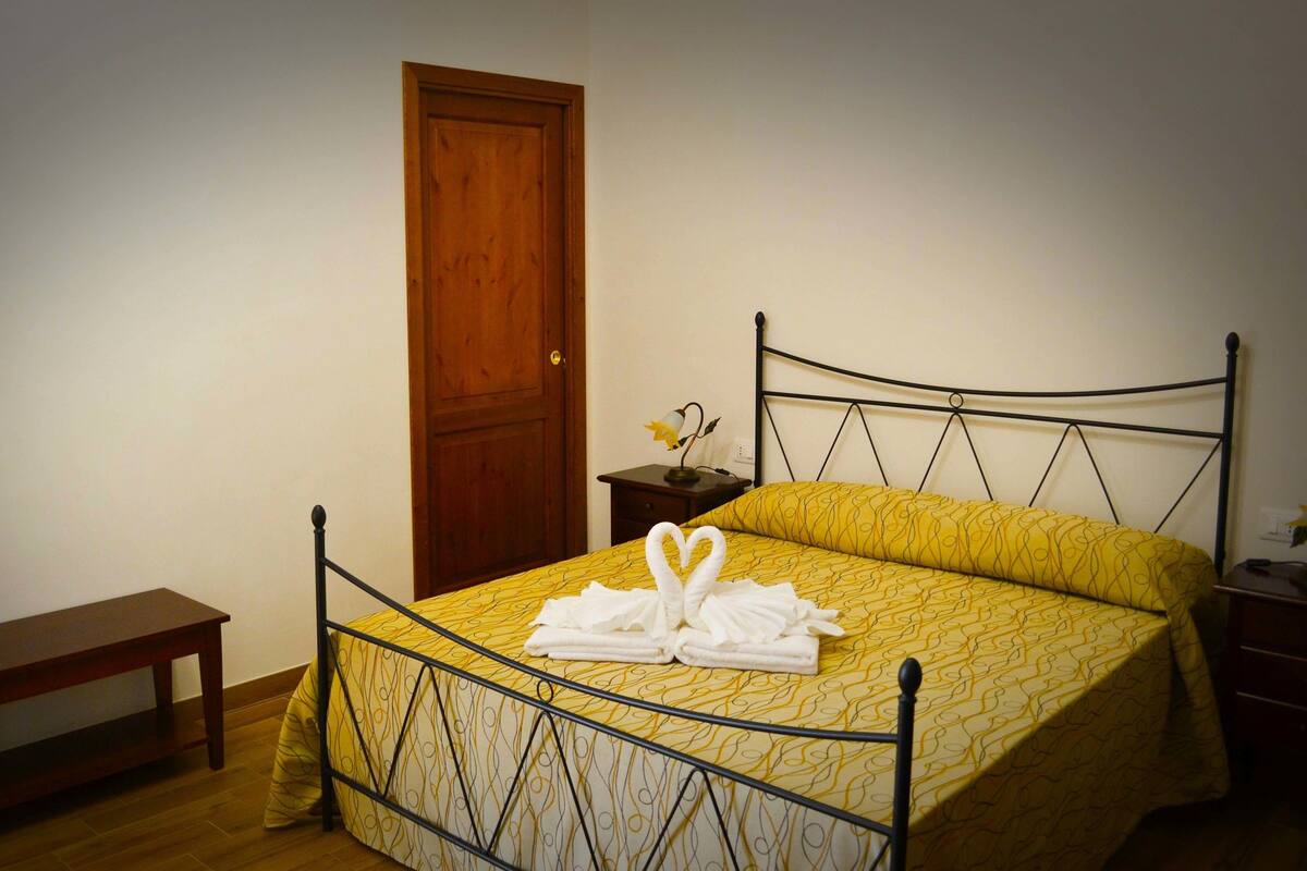 A neatly made bed with a yellow patterned bedspread, a wooden door, a small bedside table with a lamp, and a wooden bench.