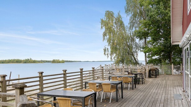 Una terraza de madera con mesas y sillas da a un lago, con una cerca de madera y árboles al fondo.