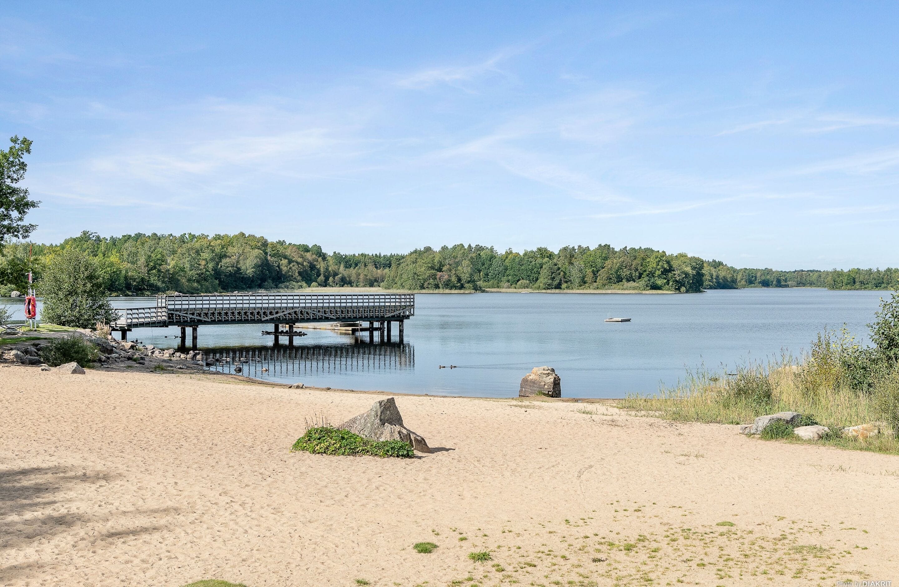 Een strand met een houten pier die zich uitstrekt over kalm water, omgeven door bomen en een heldere lucht.