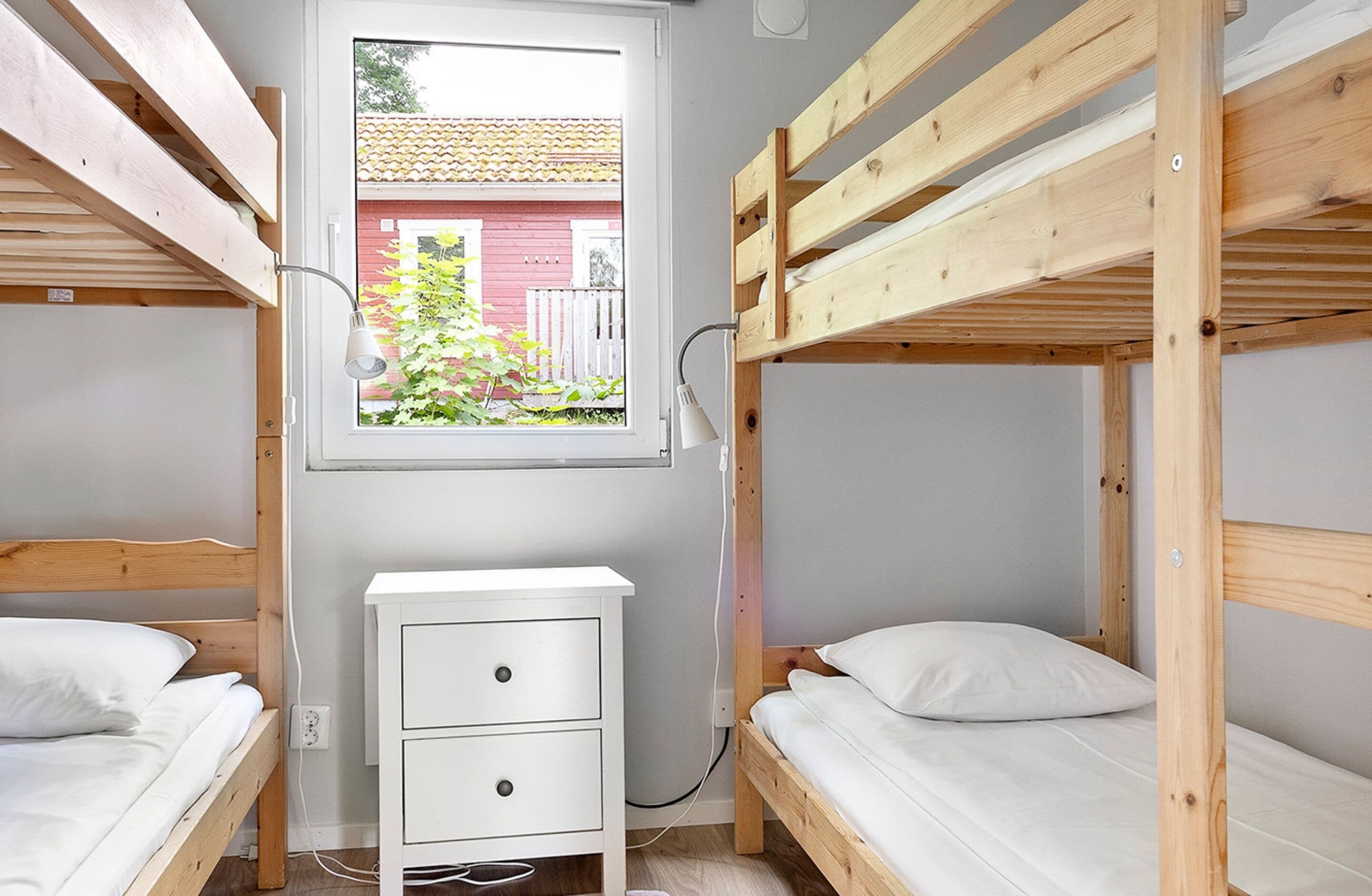 A compact room with bunk beds, a small white dresser, and a window with a view of a red building and greenery outside.