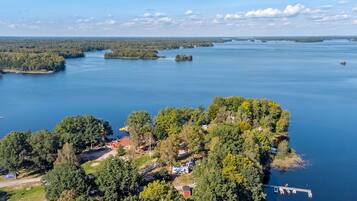 An aerial view of a lakeside property with a dock, boats, and a small house surrounded by dense trees.