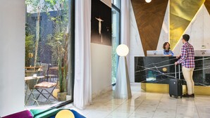 Un espace d’accueil d’hôtel au design moderne, comprenant un comptoir d’accueil, un coin salon agrémenté de coussins colorés et une vue sur l’extérieur.