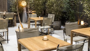Un espace café en plein air avec des tables en bois et des chaises en osier, entouré de plantes en pot et éclairé par une lumière chaleureuse.