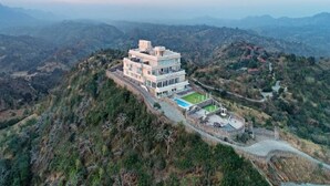 Un edificio de varios pisos con piscina y una estructura fortificada, rodeado de terreno montañoso.