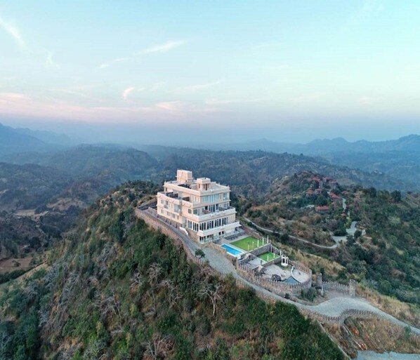 Un edificio de varios pisos con piscina y una estructura fortificada, rodeado de terreno montañoso.