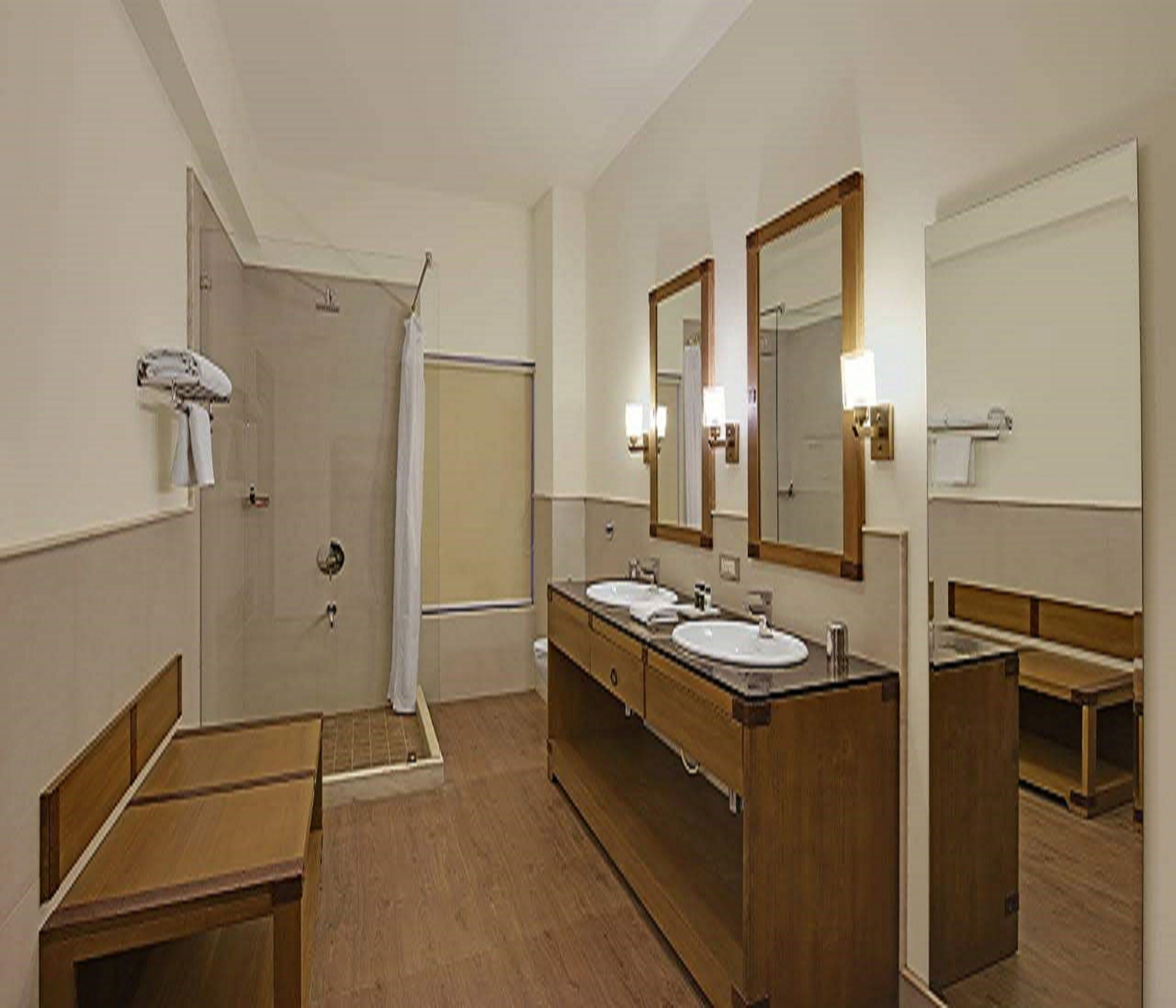 A hotel bathroom with a shower, two sinks, a mirror, and wooden benches.