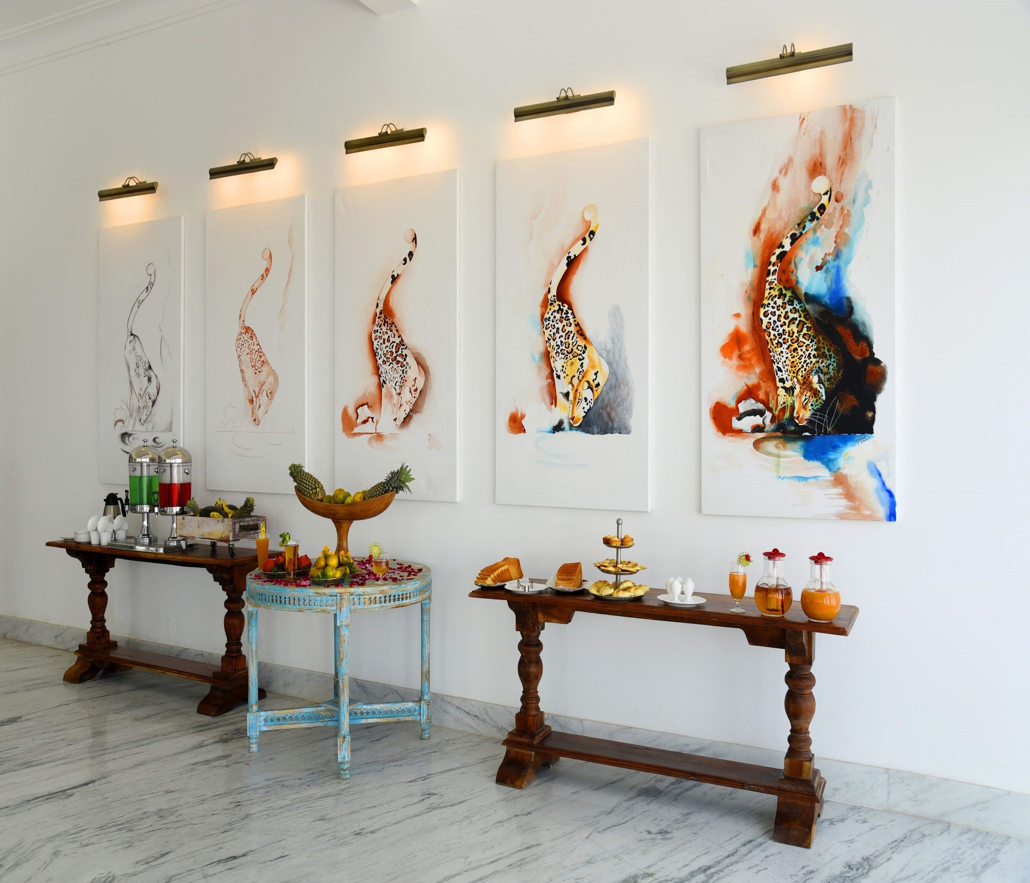 Una habitación con cuatro cuadros con motivos de jirafa en la pared, una estación de café con cafetera y tazas, y dos mesas de madera con comida y bebidas.