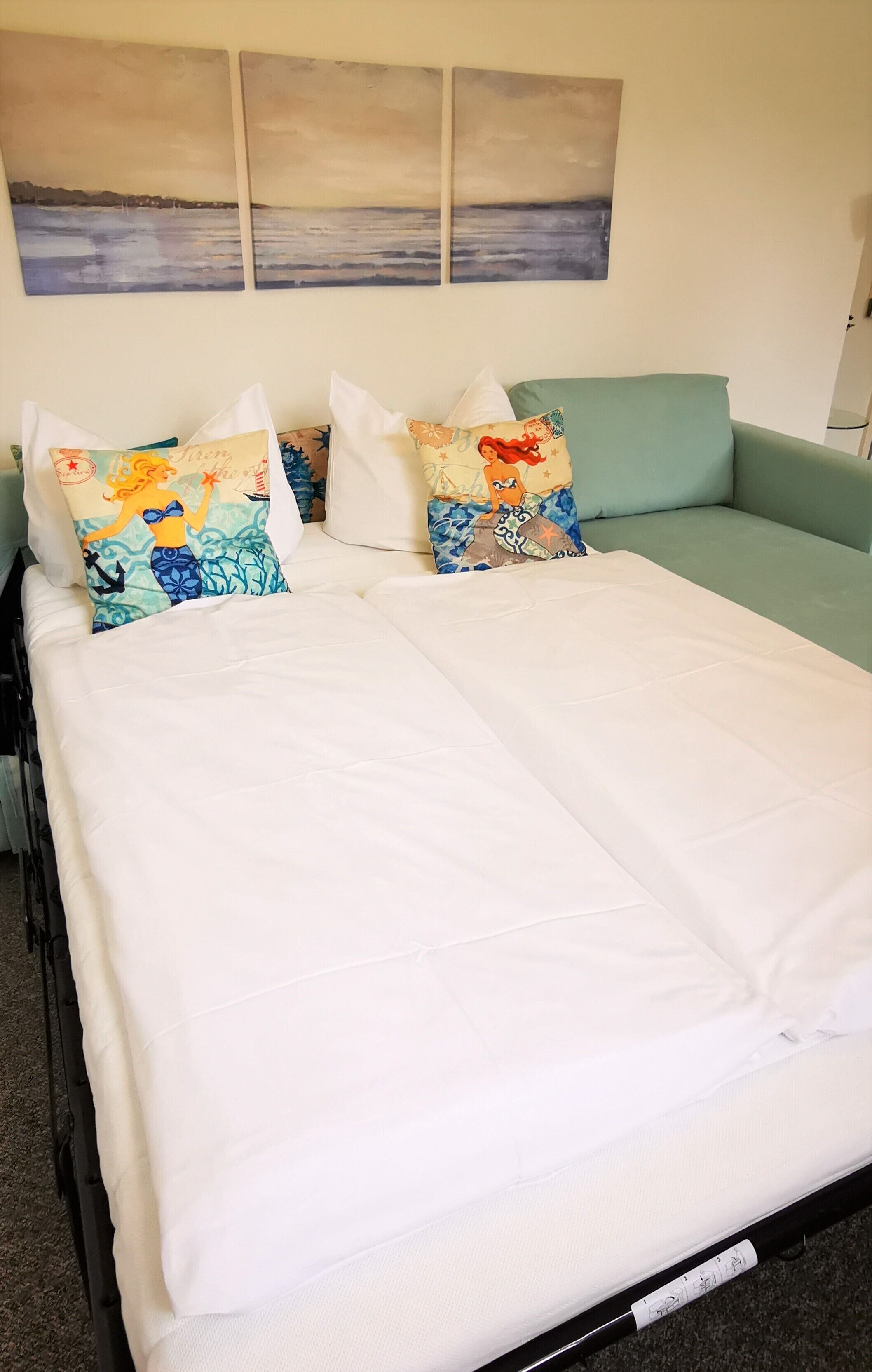 Una cama con sábanas blancas, un sofá color turquesa y tres almohadones decorativos con motivos de sirena.
