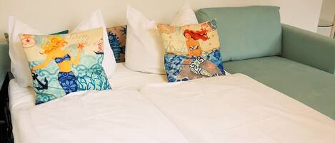 Una cama con sábanas blancas, un sofá color turquesa y tres almohadones decorativos con motivos de sirena.