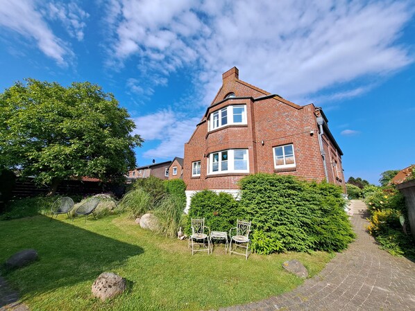 A red brick house with a large garden, a paved pathway, and outdoor seating. - Apartments mit Flair (Fehmarn)