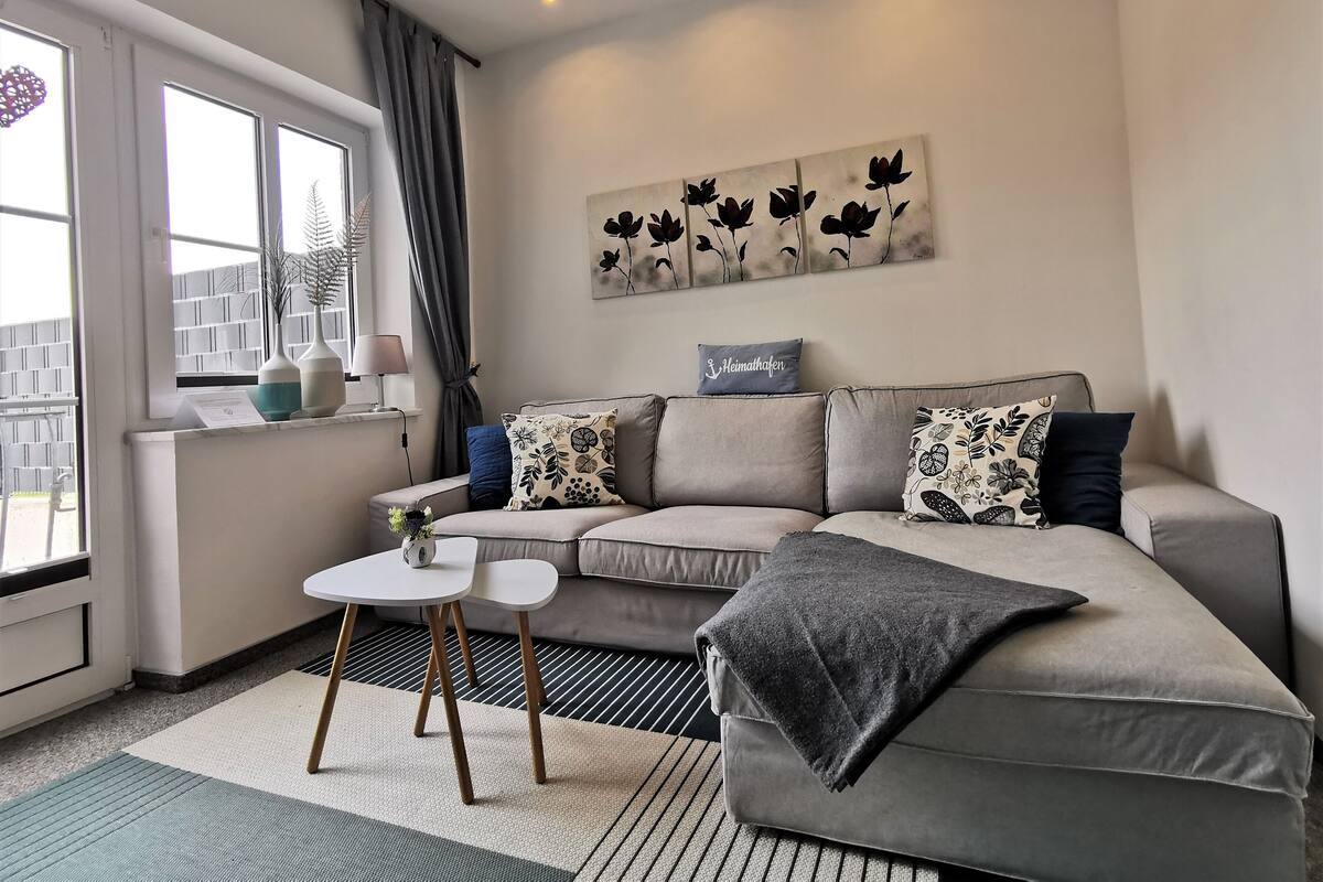 A modern living room with a grey sofa, a small white table, and a large window with curtains.