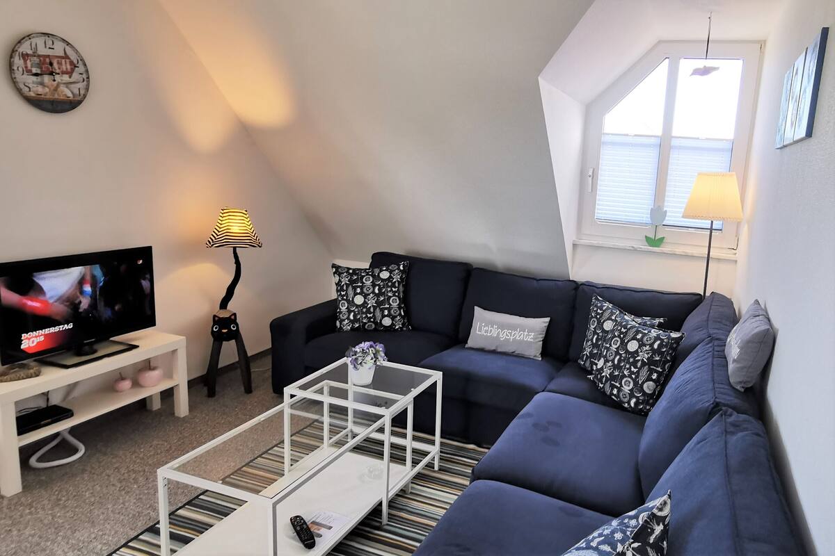 A living room with a dark blue sofa, a white coffee table, a TV on a stand, a lamp, and a window with blinds.
