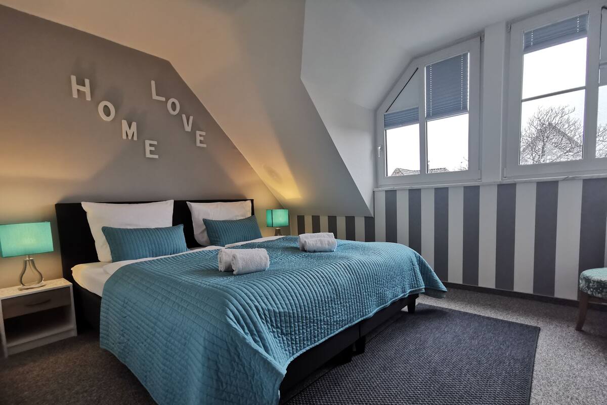 A bedroom with a bed, two bedside tables, a wall with 'HOME LOVE' letters, a window, and a striped wall.