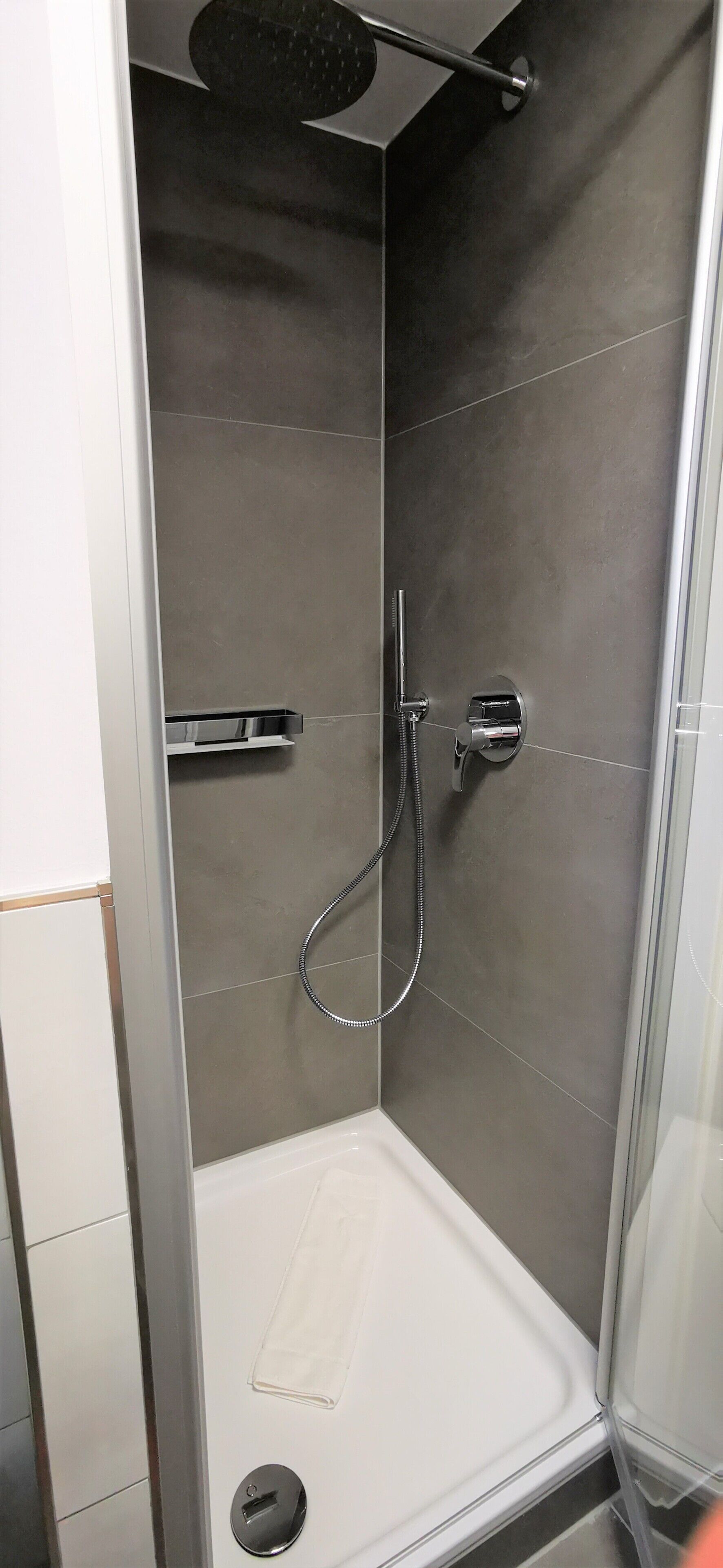 A modern shower with a handheld showerhead, a wall-mounted faucet, and a small shelf.