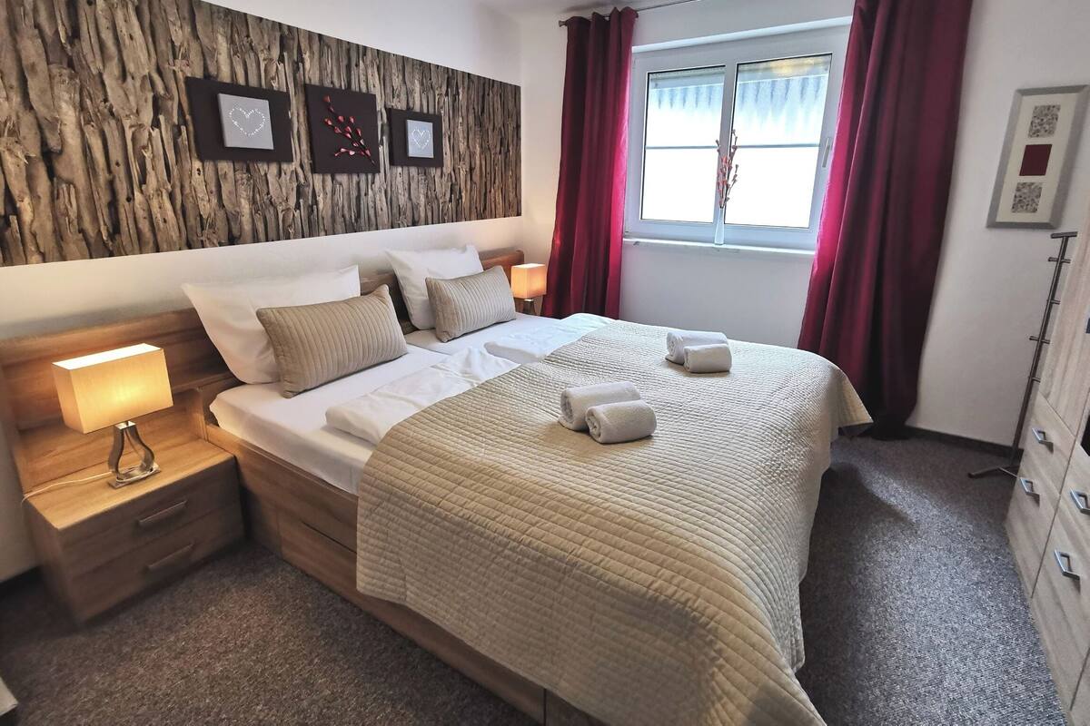 A neatly made bed with a quilted cover, two bedside lamps, a wooden headboard, and a window with red curtains.