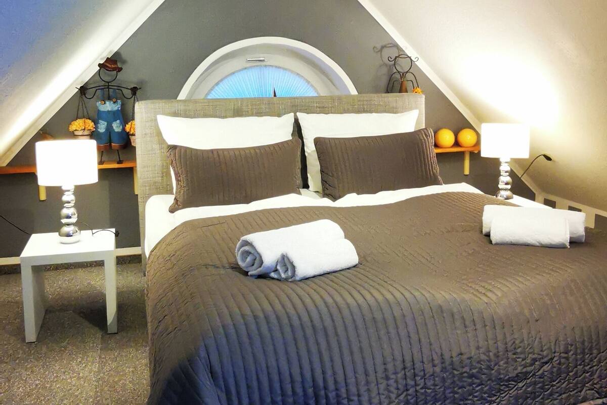 A bedroom with a large bed, two bedside tables, a lamp, and a view of the sky through a window.