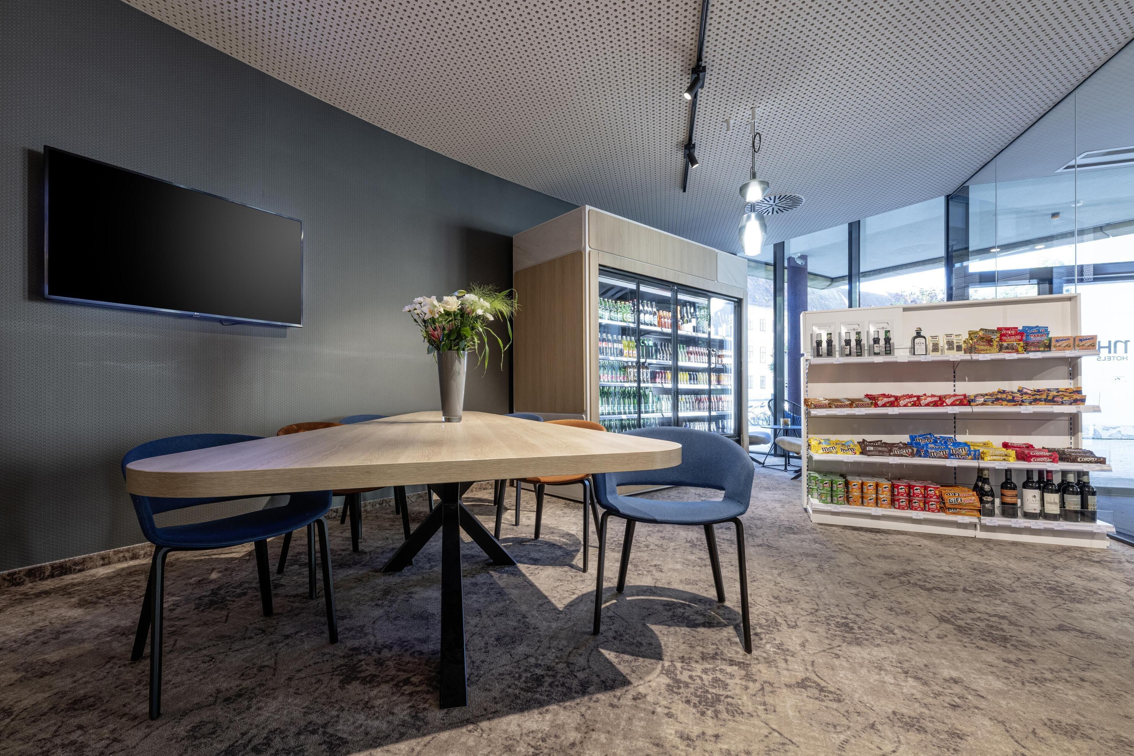A modern meeting room with a large table, chairs, a flat-screen TV, a bookshelf, and a snack shelf.