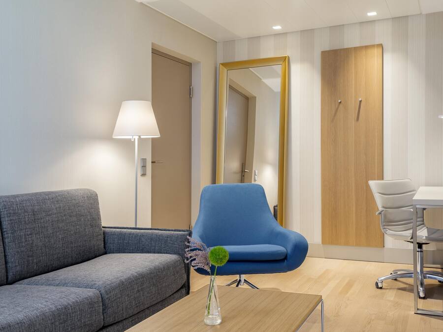 Una habitación de hotel moderna con un sofá gris, un sillón azul, un escritorio de madera y un espejo en la pared.