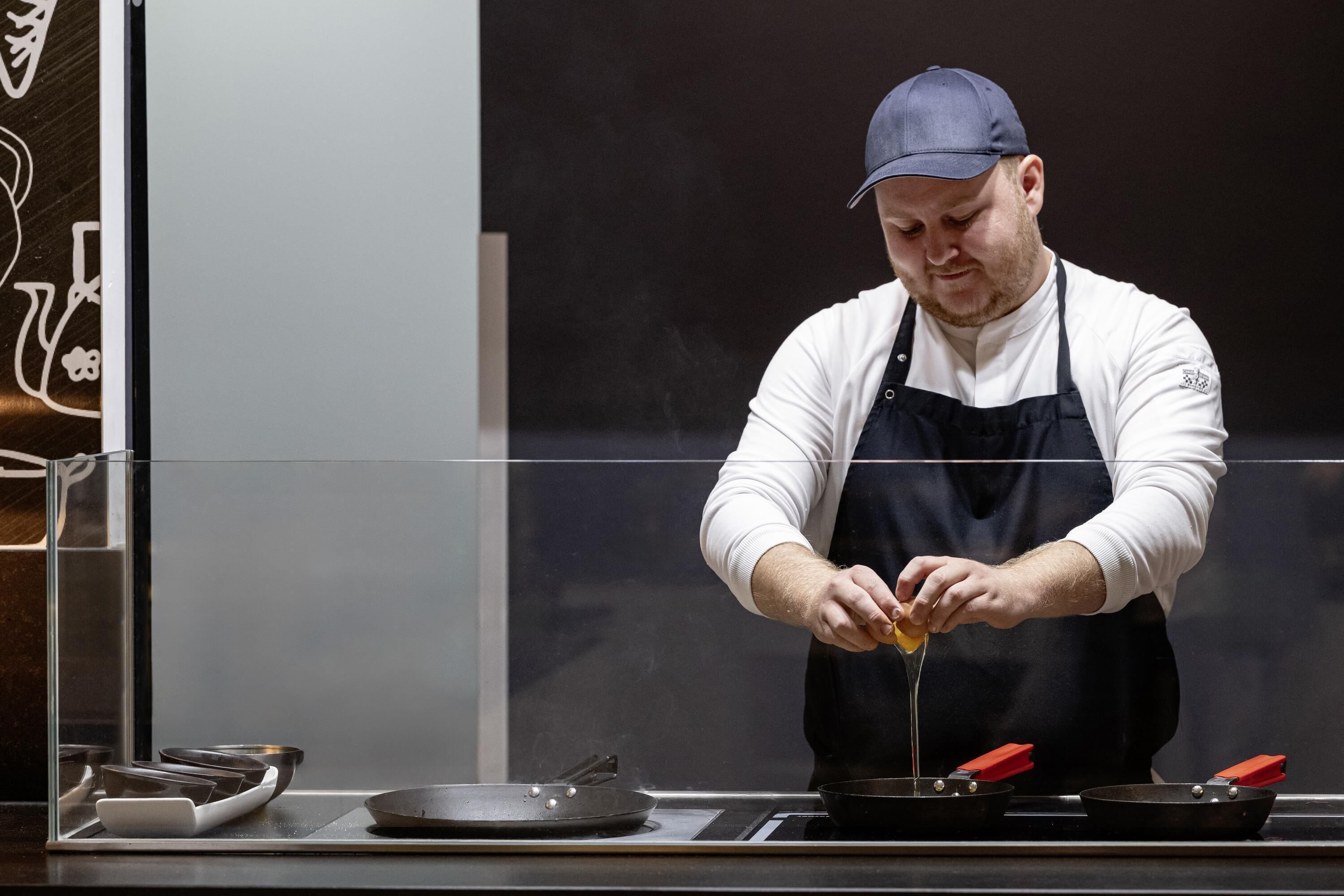 A chef in a white shirt and black apron cooking on a stove with a frying pan.