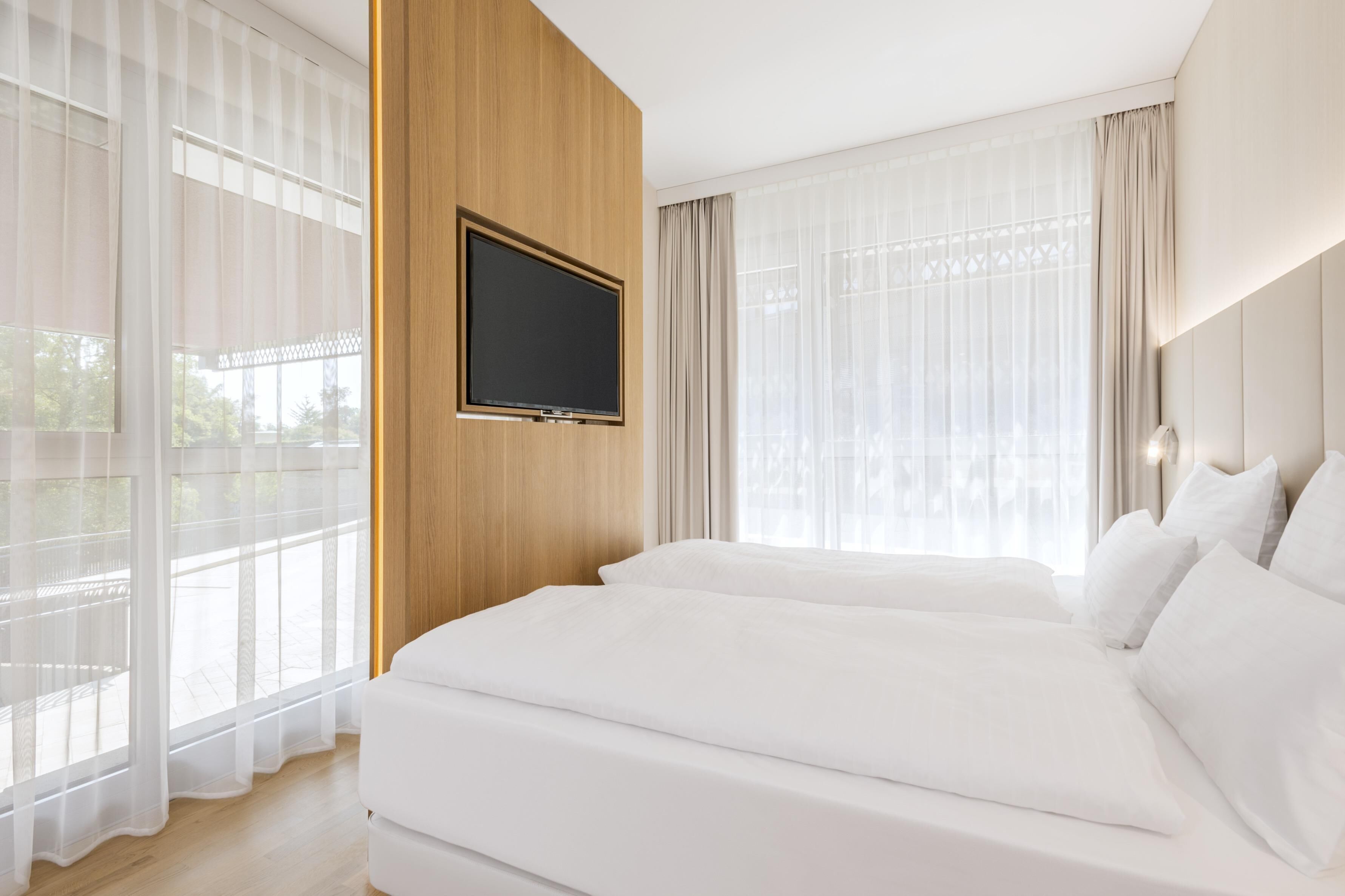 A hotel room with a large bed, a mounted TV, and large windows with sheer curtains.
