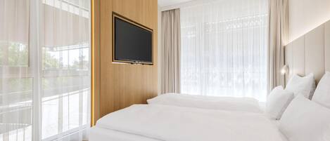 A hotel room with a large bed, a mounted TV, and large windows with sheer curtains.