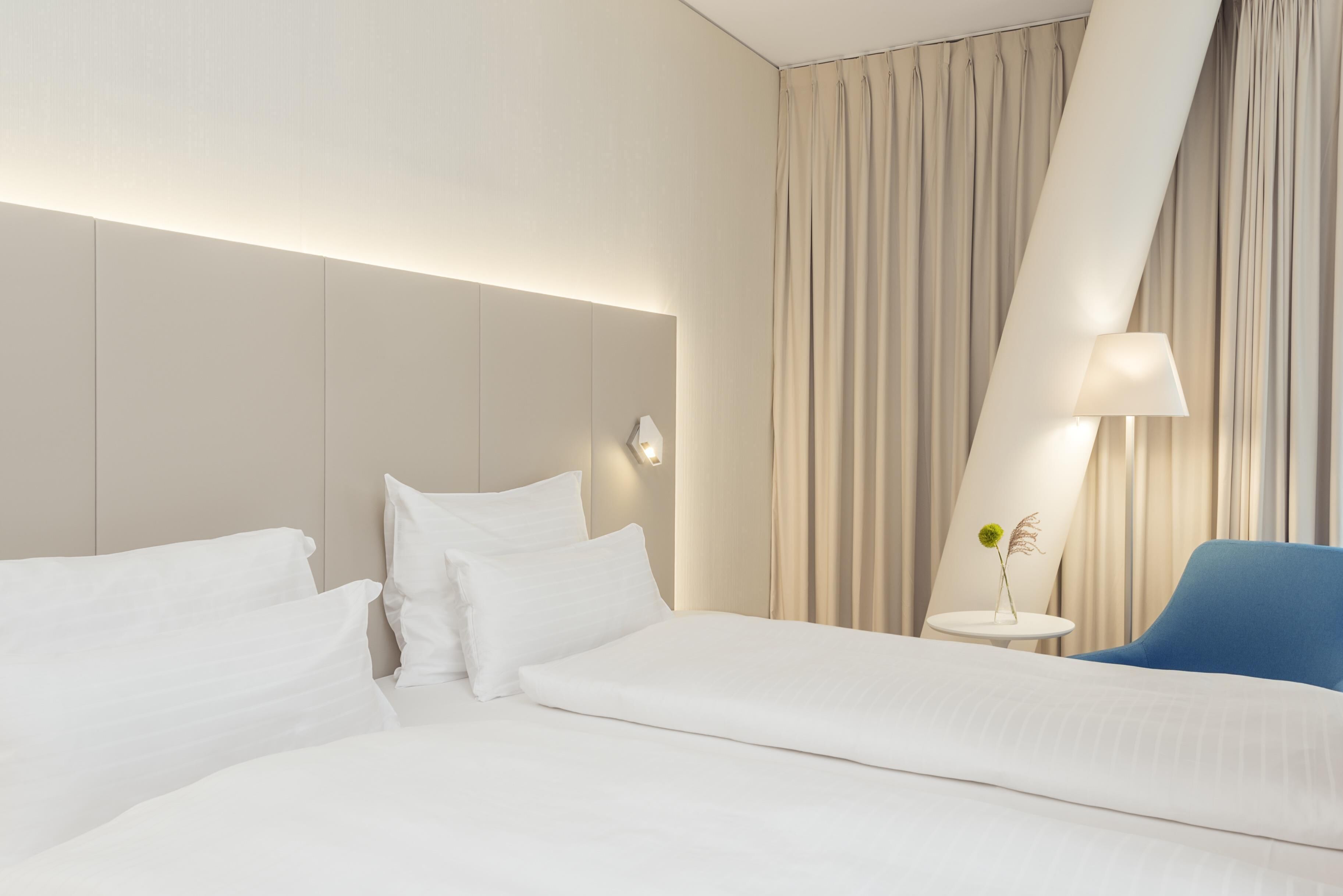 A modern hotel room with a large bed, a blue chair, a small table with a vase, and a large window with curtains.