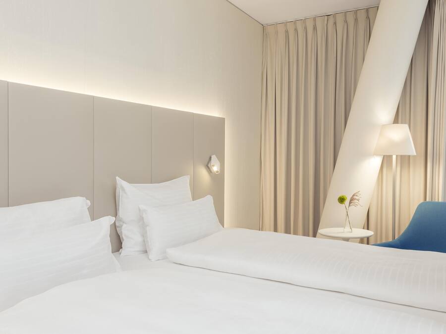 Habitación de hotel moderna con una cama grande, una silla azul, una mesita con un florero y un ventanal con cortinas.