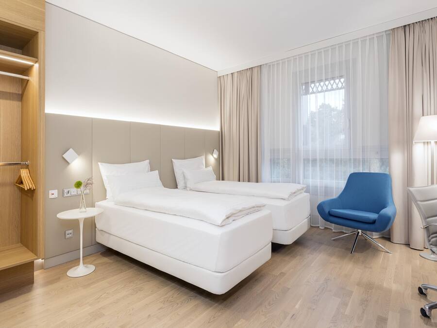 Une chambre d’hôtel moderne avec deux lits, un bureau, une chaise et une grande fenêtre avec des rideaux.