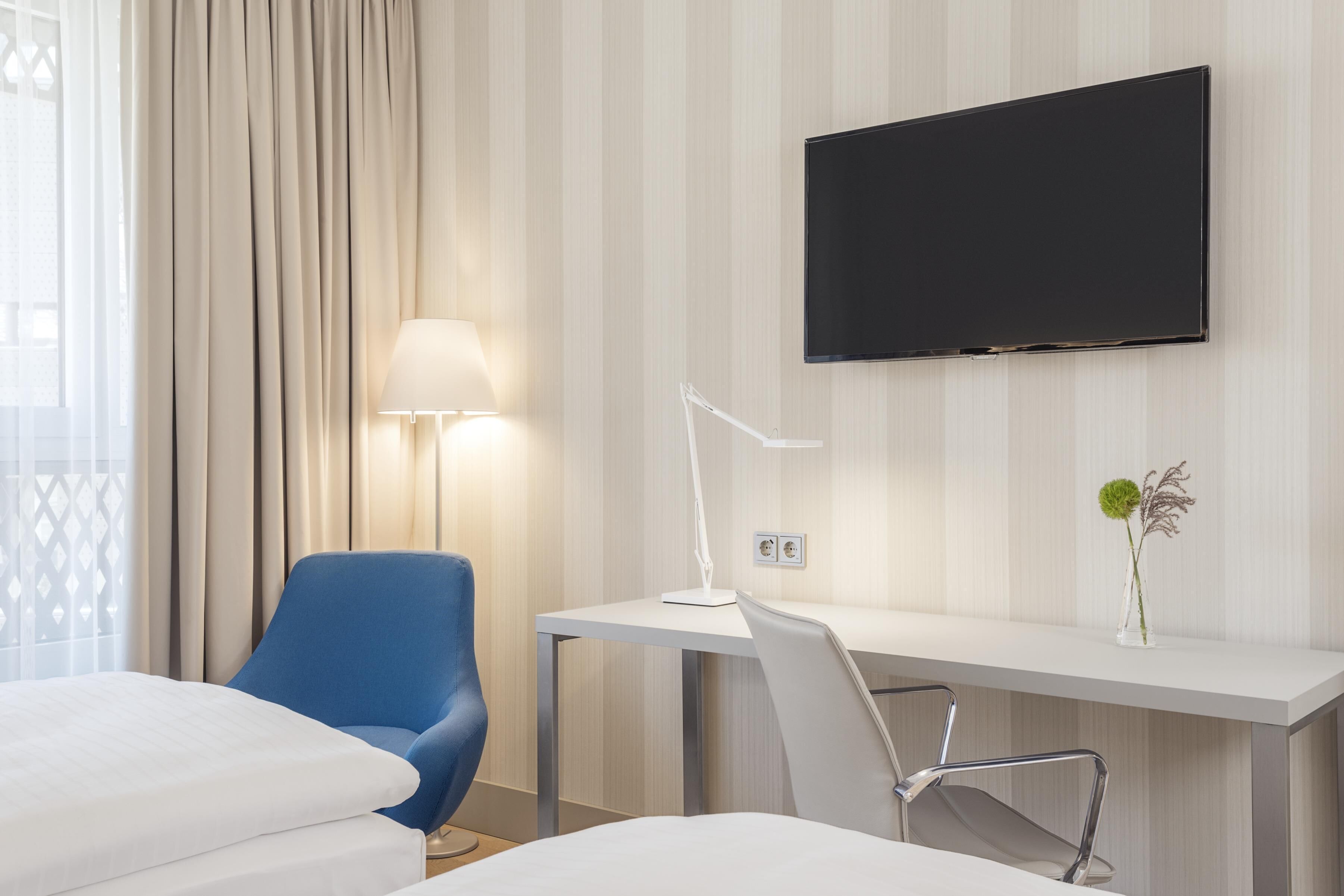 Habitación de hotel con cama, escritorio, silla, televisor, lámpara y un florero con planta.