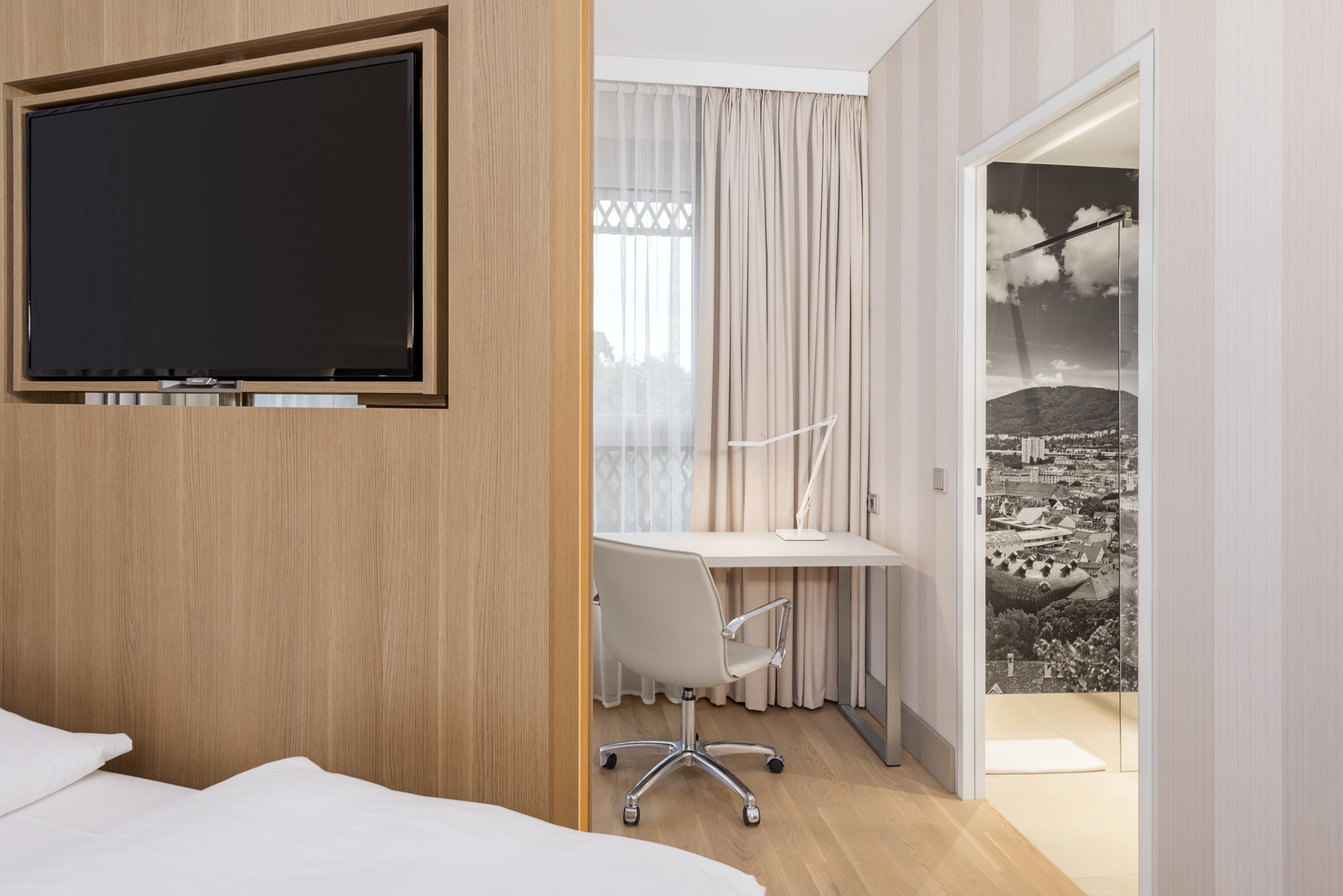 Habitación de hotel con cama, escritorio con silla, televisor y vista a un paisaje urbano a través de la ventana.
