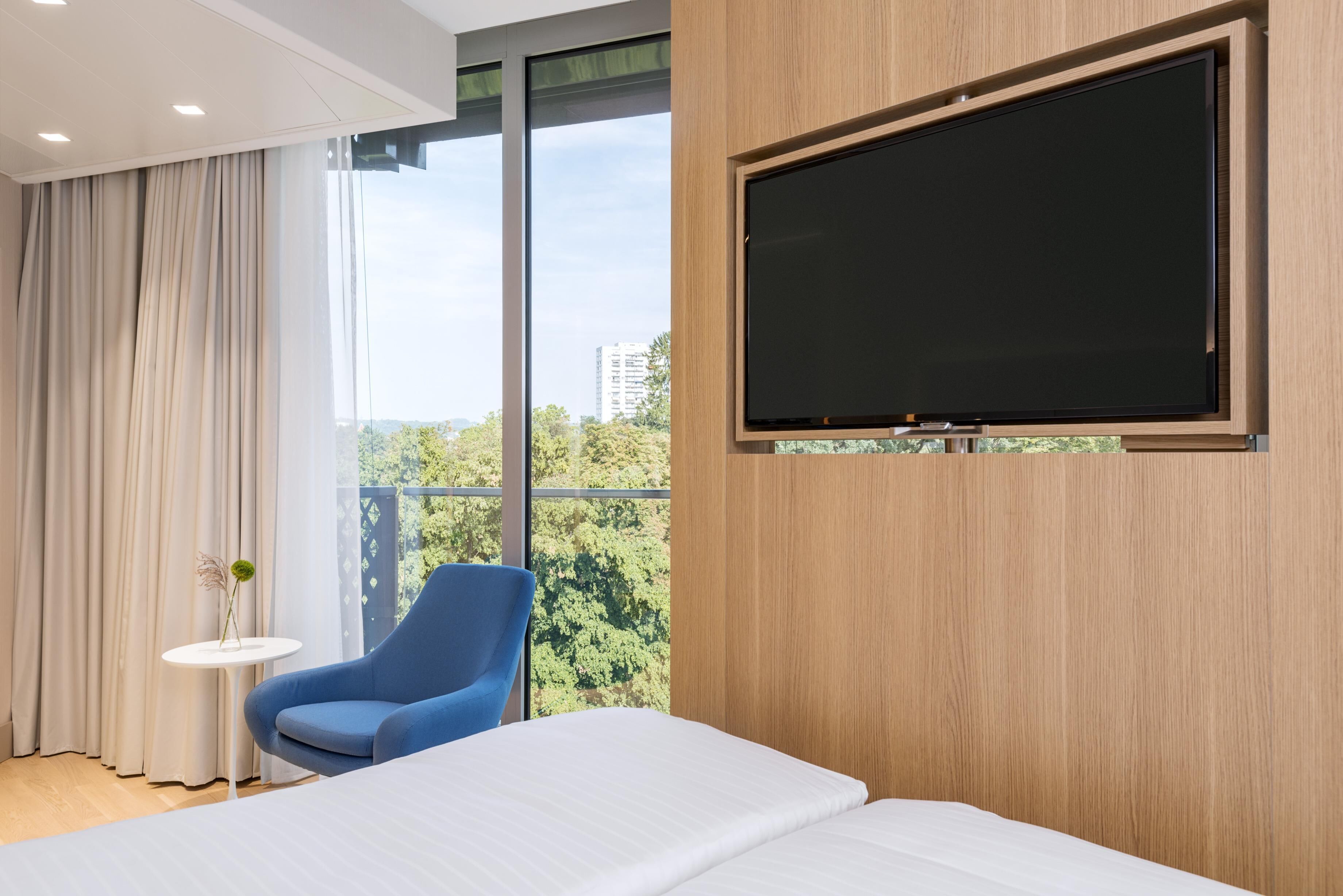 Una habitación de hotel moderna con un ventanal grande, un televisor de pantalla plana montado en la pared, una silla azul, una mesita redonda con un florero y una cama bien hecha.