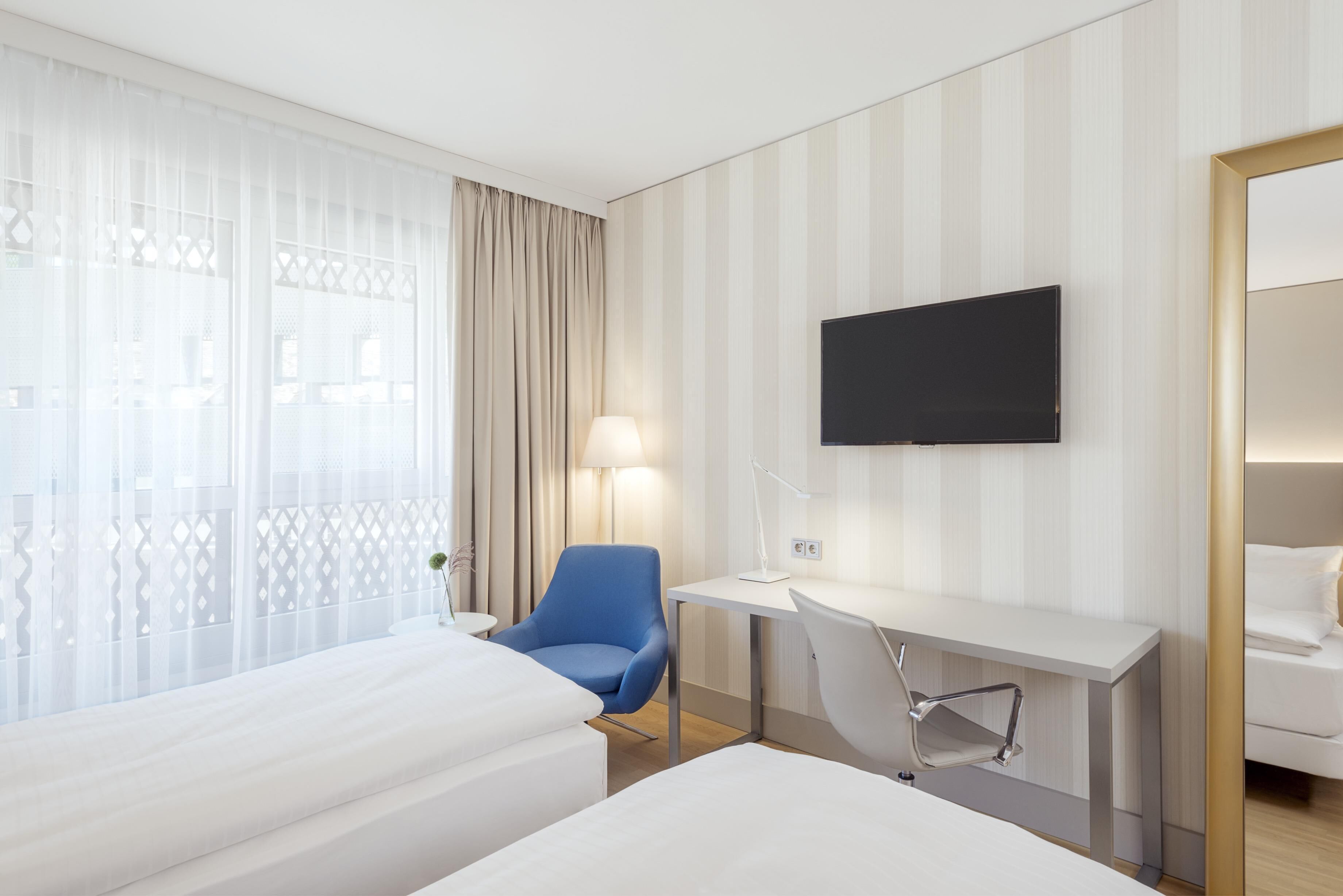 A hotel room with two beds, a desk, a chair, a TV, and a window with curtains.