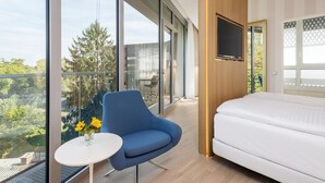 Ein modernes Hotelzimmer mit großem Fenster, einem blauen Sessel, einem weißen Bett und einem kleinen Tisch mit Blumen.