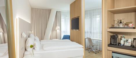 A modern hotel room with a bed, a desk, a chair, a TV, and a minibar.