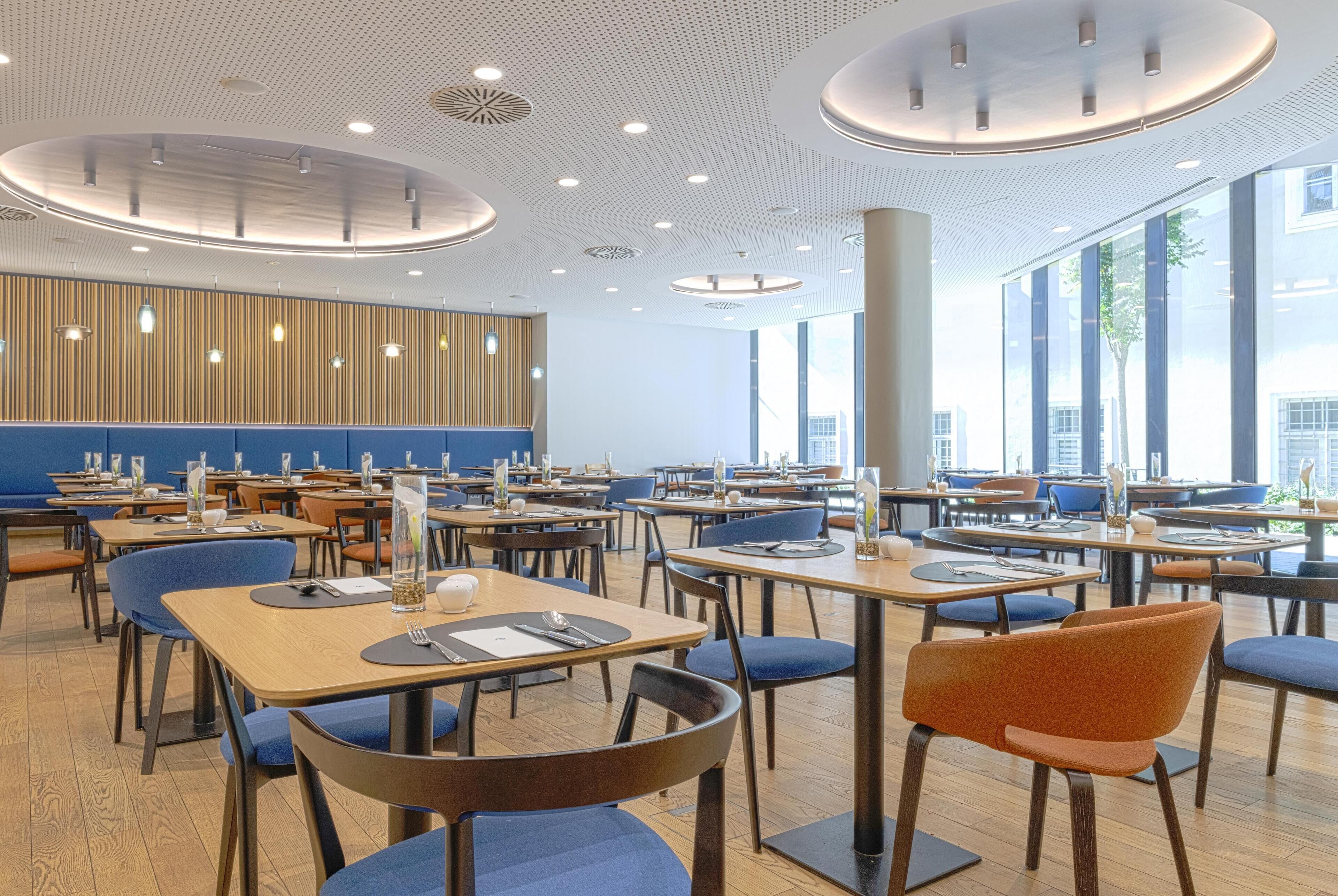Un espace repas moderne doté de plusieurs tables et chaises, de grandes fenêtres et d’un mur revêtu de panneaux en bois.