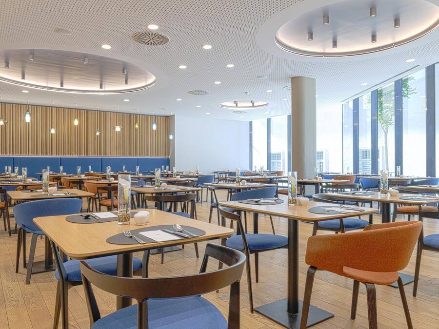 A modern dining area with multiple tables and chairs, large windows, and a wooden panel wall.