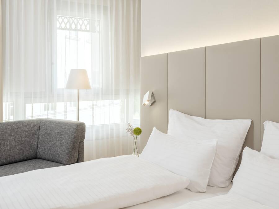 Habitación de hotel con cama, sofá gris, lámpara y un florero con una sola flor.
