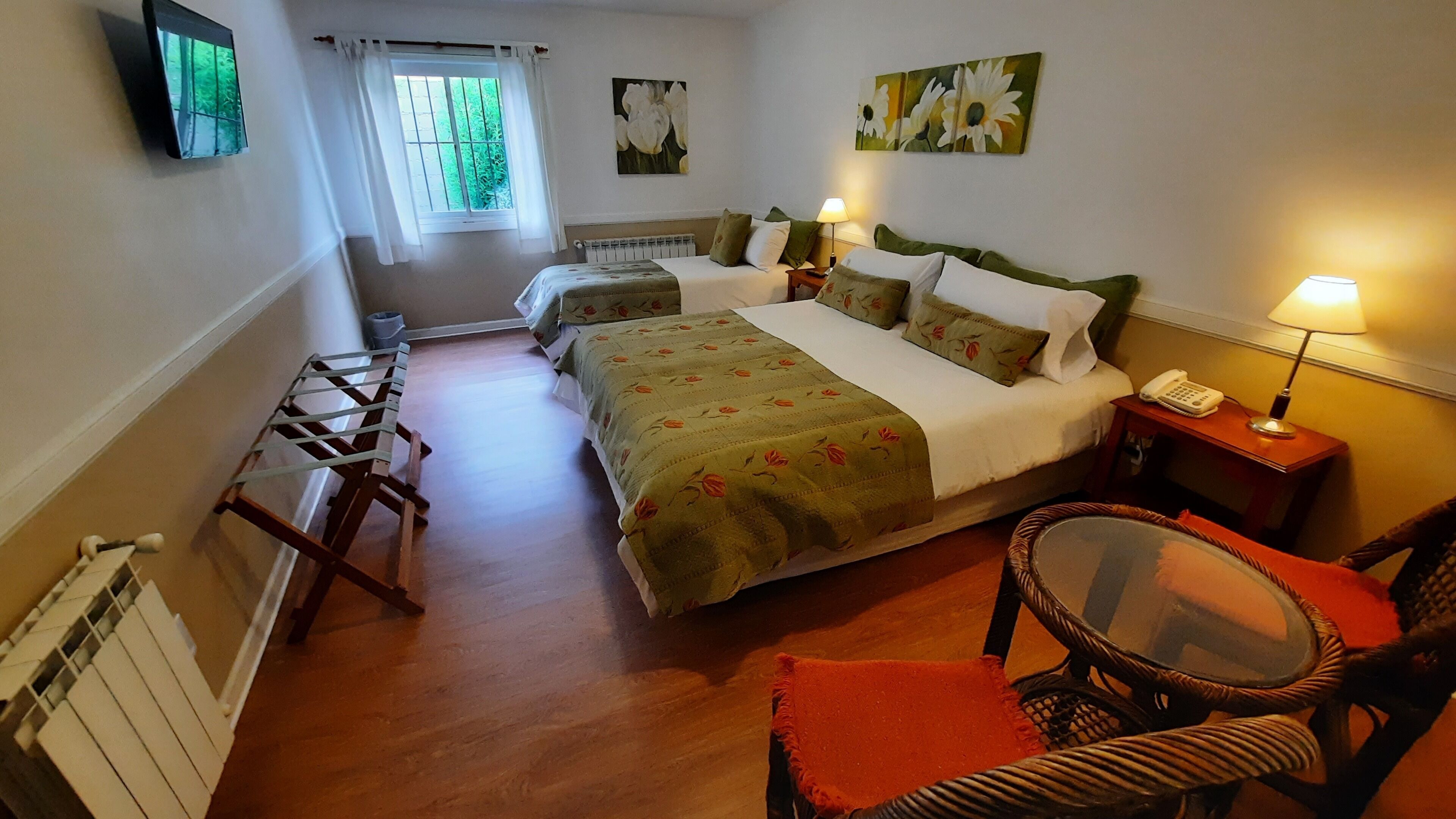 Una habitación de hotel con cama, una silla, una mesa, un radiador, una ventana con cortinas y obras de arte en las paredes.