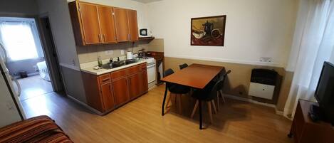 A compact kitchen with wooden cabinets, a dining table with chairs, a microwave, and a painting on the wall.