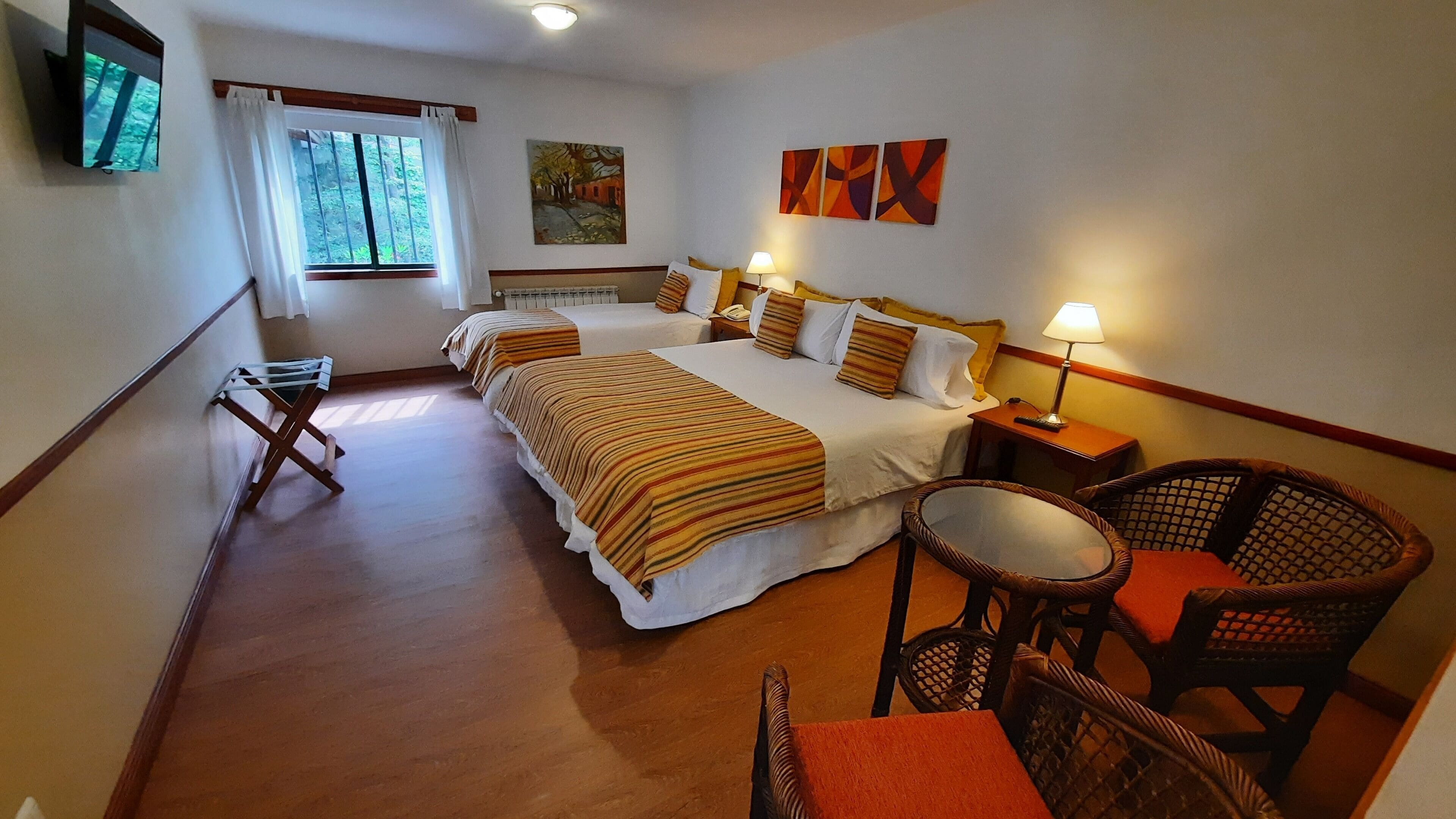 Hotellihuone, jossa on kaksi sänkyä, televisio, ikkuna verhoilla, taulu ja pieni pöytä tuolilla.